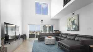A modern living room with a large flat-screen TV, a sectional sofa, a round coffee table, and a large window offering a city view. - Portside Villas Unit 6 (Pensacola Beach)