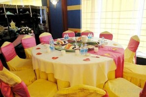 Ein runder Tisch mit weißer Tischdecke, umringt von gelben Stühlen mit rosa Bändern, gedeckt für eine Mahlzeit mit verschiedenen Speisen und Getränken.