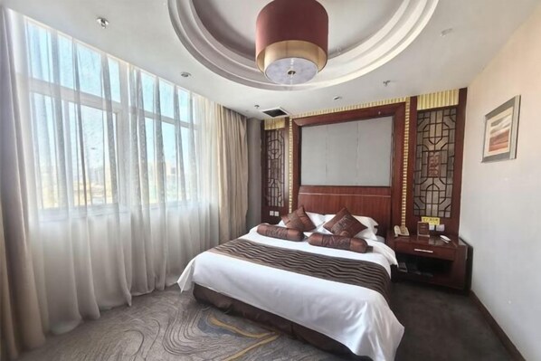 Une chambre d’hôtel avec un grand lit, un bureau, une chaise, une fenêtre avec des rideaux et un luminaire au plafond.