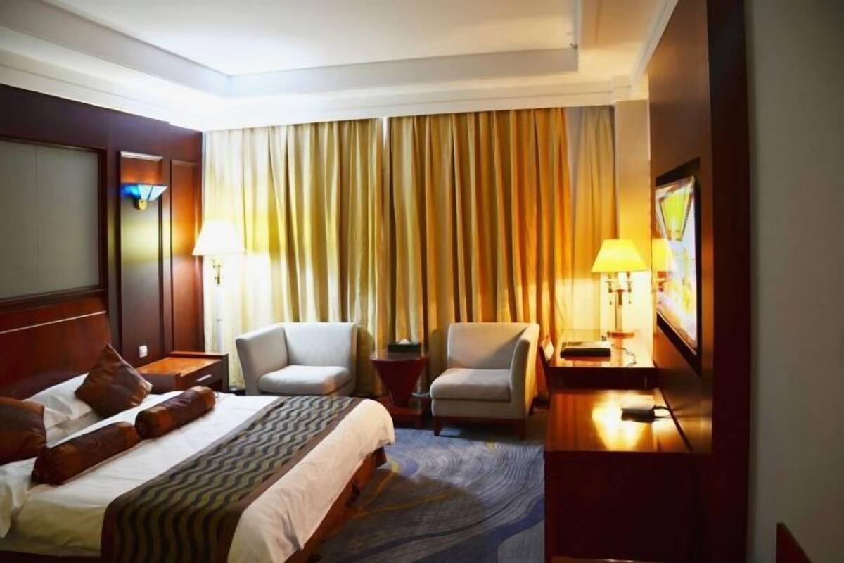 Une chambre d’hôtel comprenant un lit, deux fauteuils, un bureau avec une lampe et une grande fenêtre avec des rideaux.