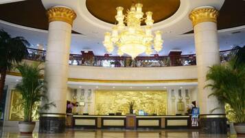 A hotel lobby with a grand chandelier, a reception desk, and a balcony with a railing.