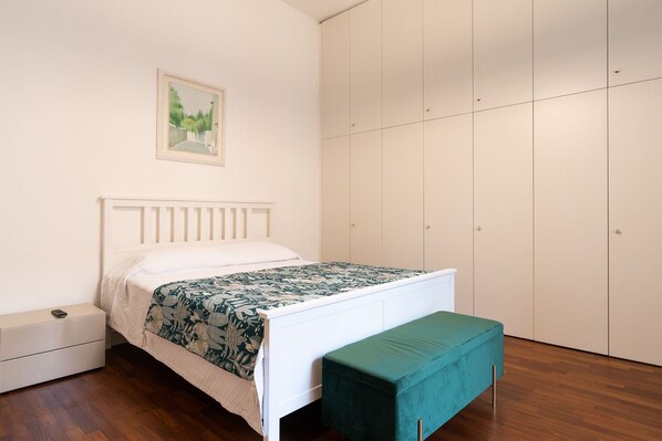 Un dormitorio con cama, mesita de noche, banco, armario y un cuadro en la pared.