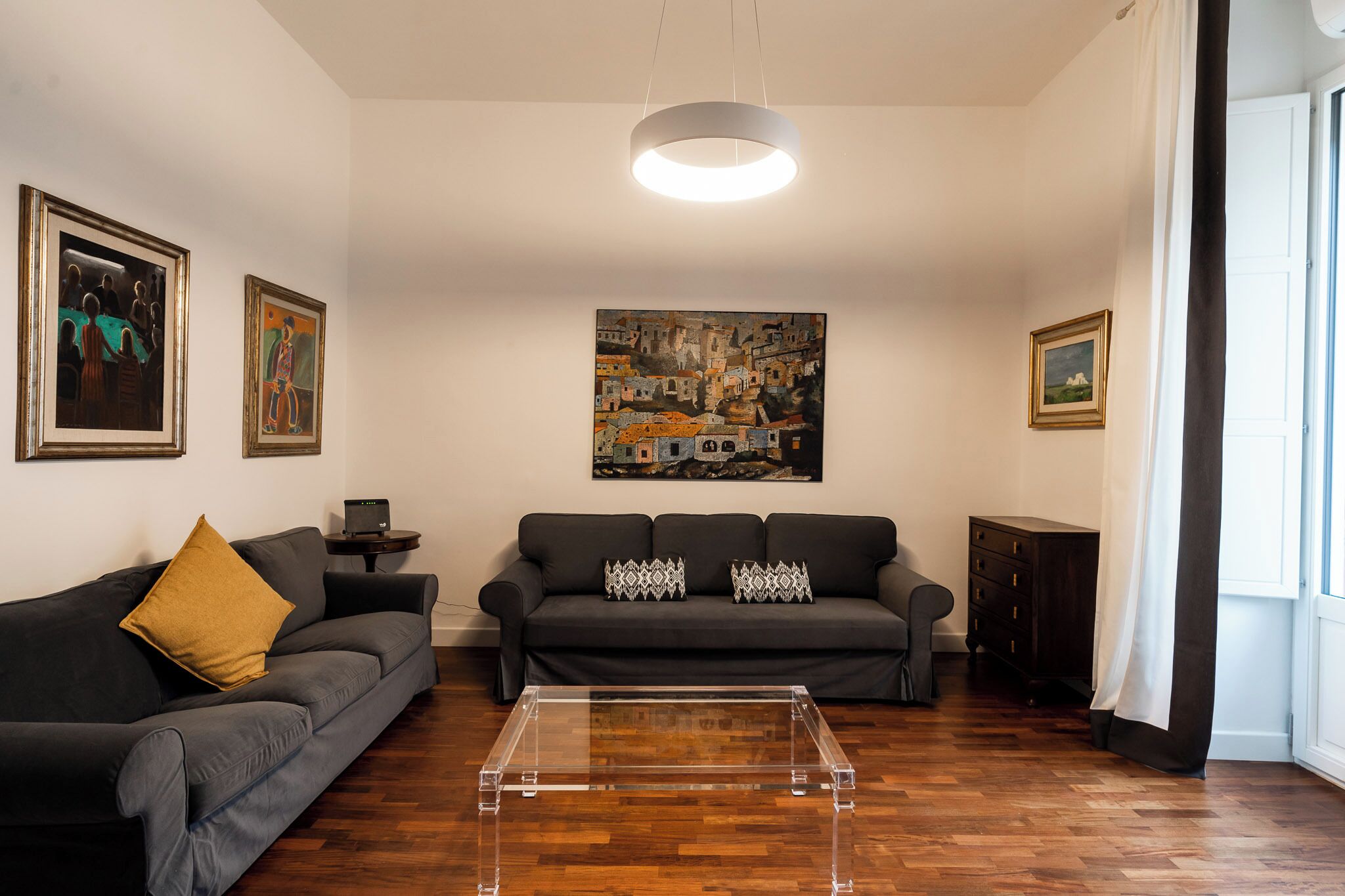 A living room with a dark grey sofa, a clear glass coffee table, and a large painting on the wall.