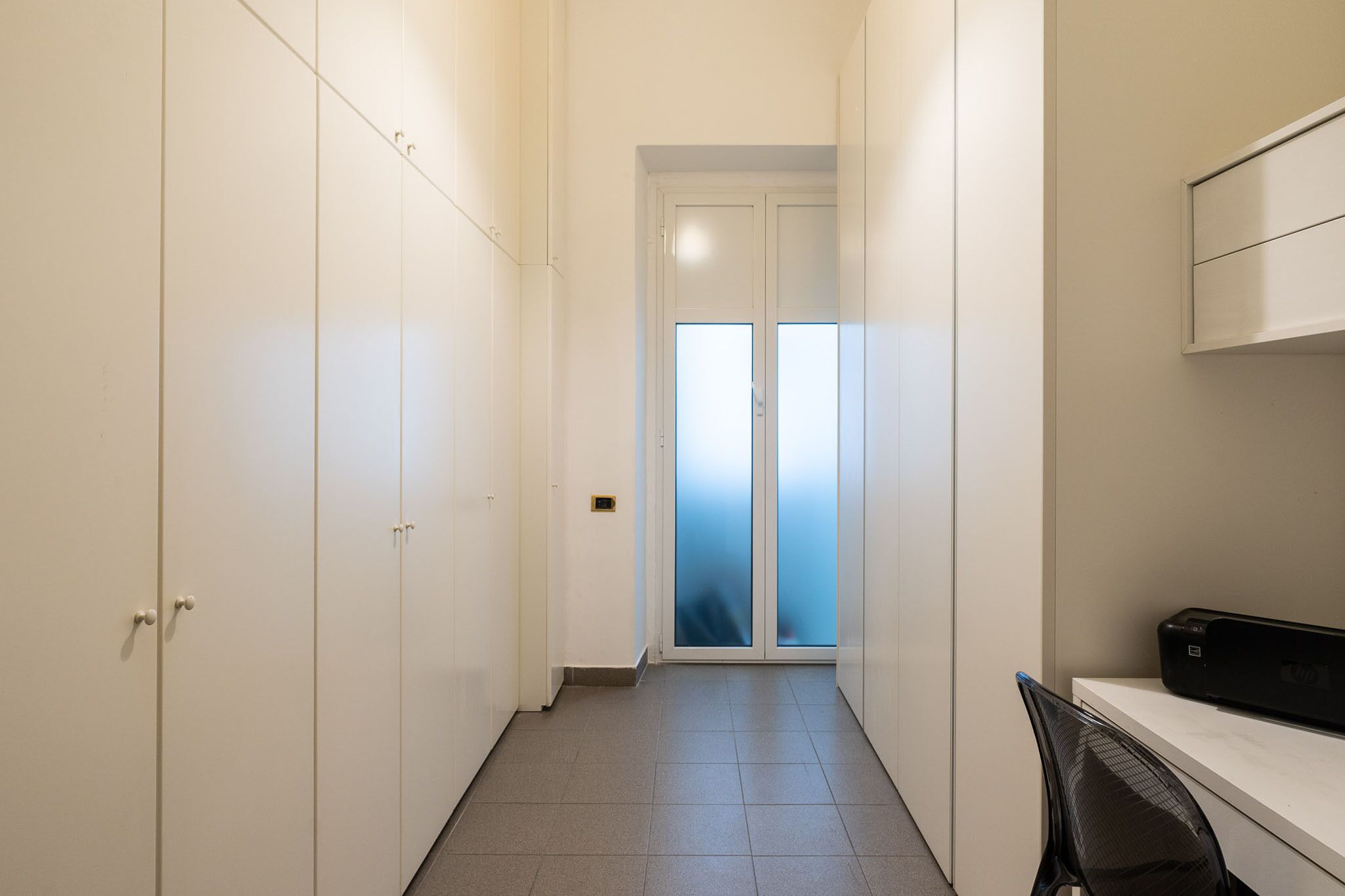 A narrow hallway with white cabinets, a door with frosted glass panels, and a desk with a printer.