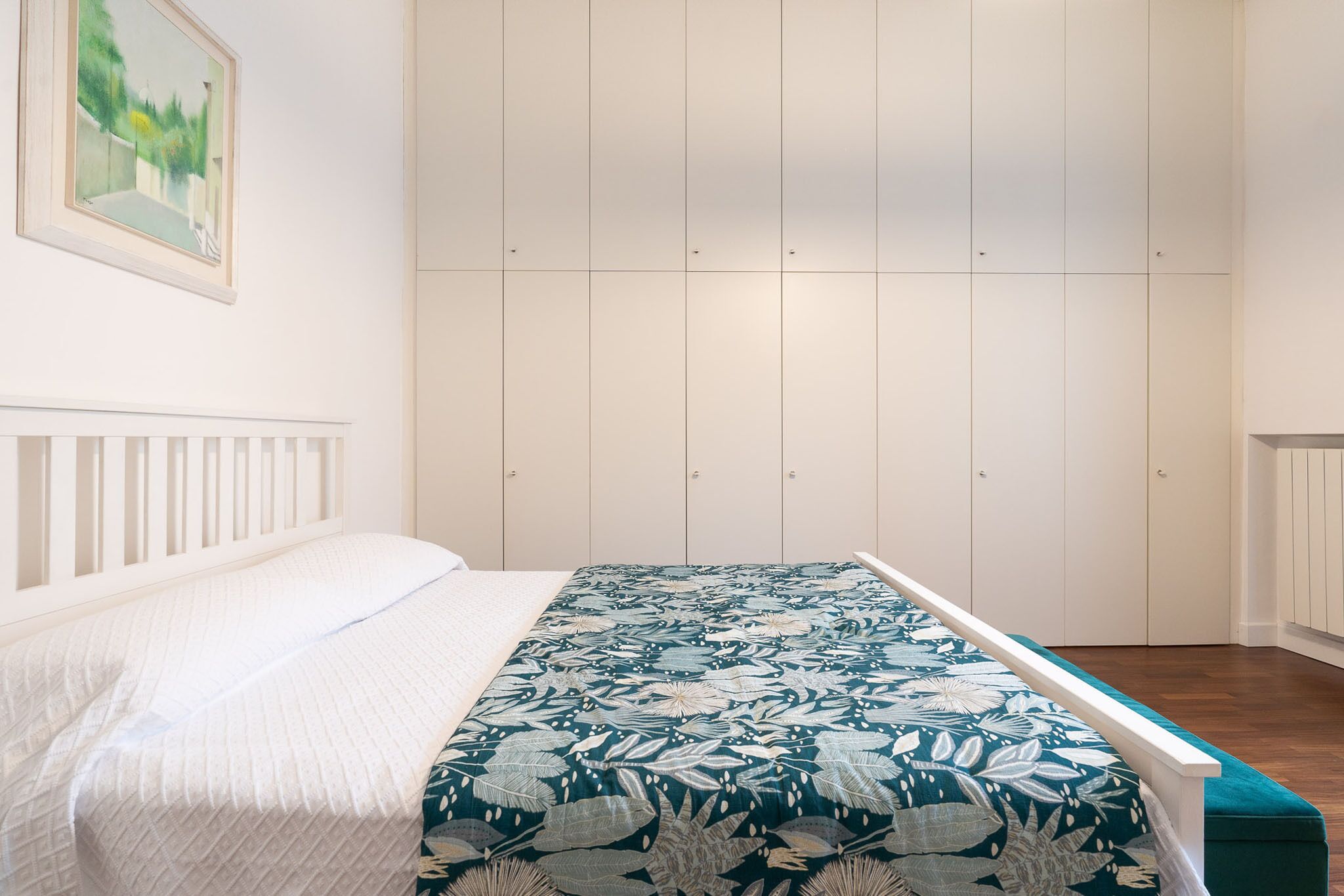 A bedroom with a bed, a white headboard, a large window, and a wardrobe with multiple doors.