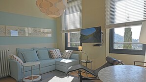 A modern living room with a sectional sofa, a round table, and a reclining chair. There are two windows with blinds, a flat-screen TV mounted on the wall, and a radiator. - VILLA ALFONSA, Apartment "LA TERRASSE" exceptional sea view, 1st floor. (Saint-Raphaël Agay)