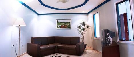 A living room with a brown sectional sofa, a standing lamp, a ceiling fan, a framed landscape painting, and a window with a view.