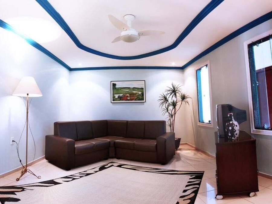 A living room with a brown sectional sofa, a standing lamp, a ceiling fan, a framed landscape painting, and a window with a view.