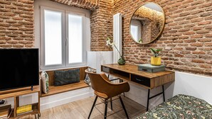 A room with a brick wall, a large window, a desk with a chair, a television, and a bed with a floral bedspread.