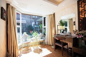 A hotel room with a large window, a desk with a chair, a small table, and a plant.