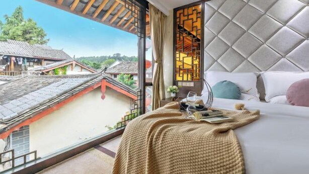 A bedroom with a large window, a bed with a white headboard, and a view of traditional buildings outside. - Weaving Sunshine Vacation Designer Ancient City View Garden Hotel (the Old Town of Lijiang) (Lijiang)