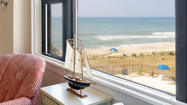 Quarto com vista para a praia, um modelo de veleiro e uma poltrona estampada.