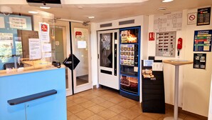 Zona de recepción de un hotel con un mostrador azul, un teléfono público, un quiosco de autoservicio y varios carteles informativos en las paredes.