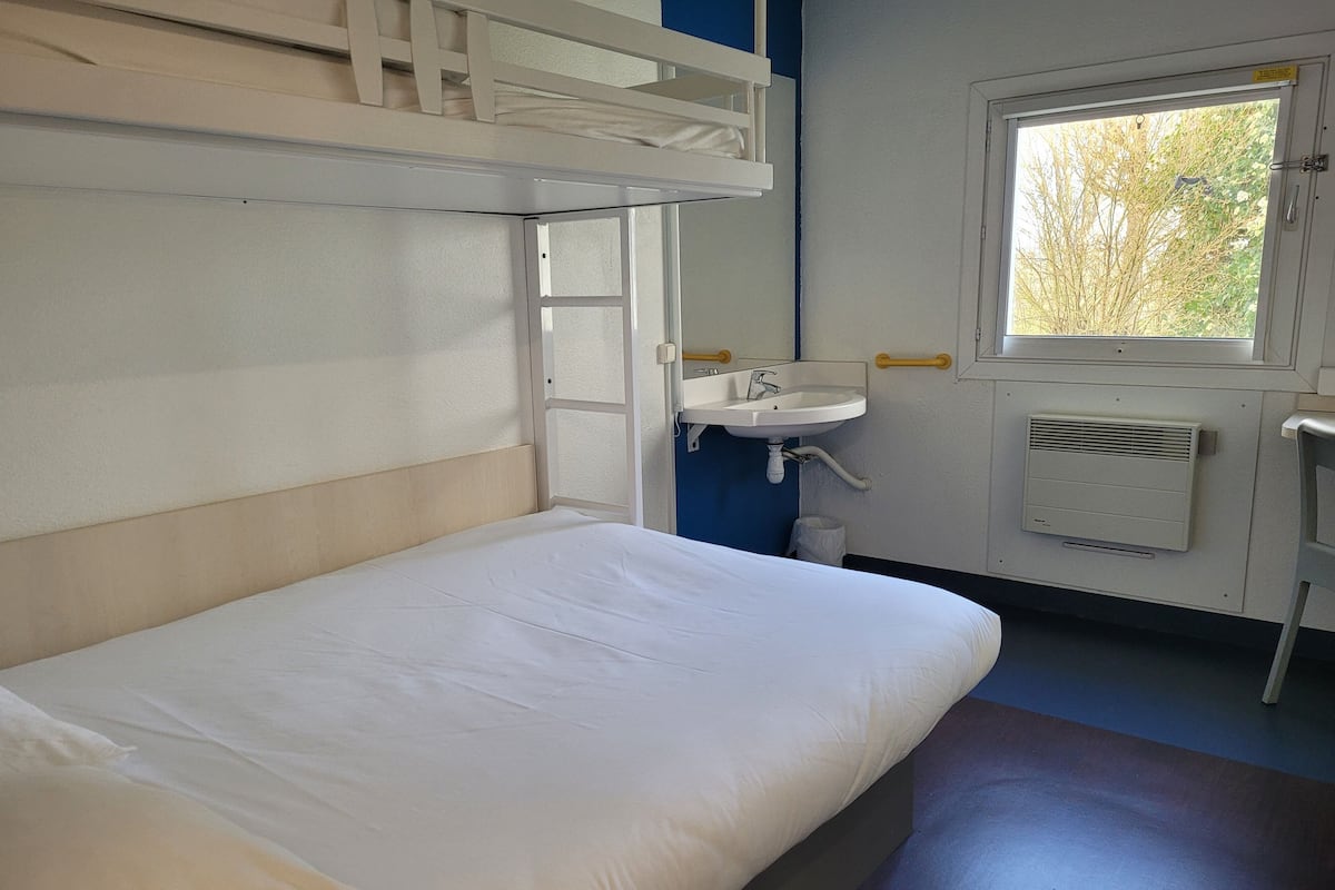 Una cama litera con colchón y almohada blancos, un lavamanos blanco con espejo encima y una ventana con vistas a árboles.