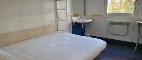 Una cama litera con colchón y almohada blancos, un lavamanos blanco con espejo encima y una ventana con vistas a árboles.