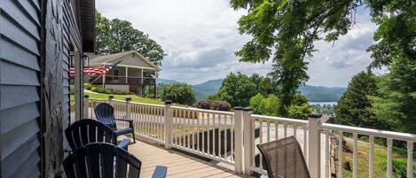 Una terraza de madera con mobiliario exterior, barandilla blanca y vista a una casa y montañas al fondo.