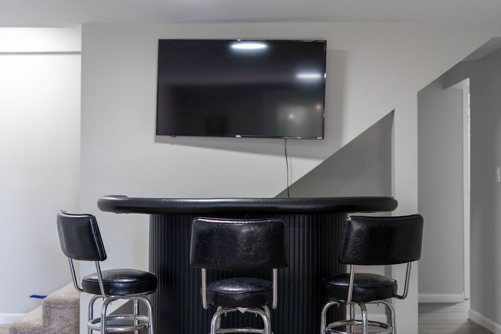 Ein moderner Barbereich mit Wand-Fernseher, drei schwarzen Barhocker und einer Treppe im Hintergrund.