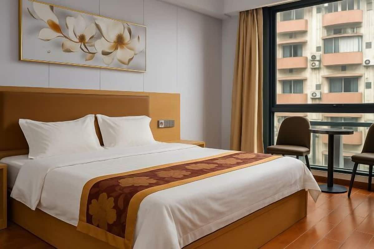 Habitación de hotel con cama, mesita de noche, una silla, una pequeña mesa y ventana con vista a edificios.