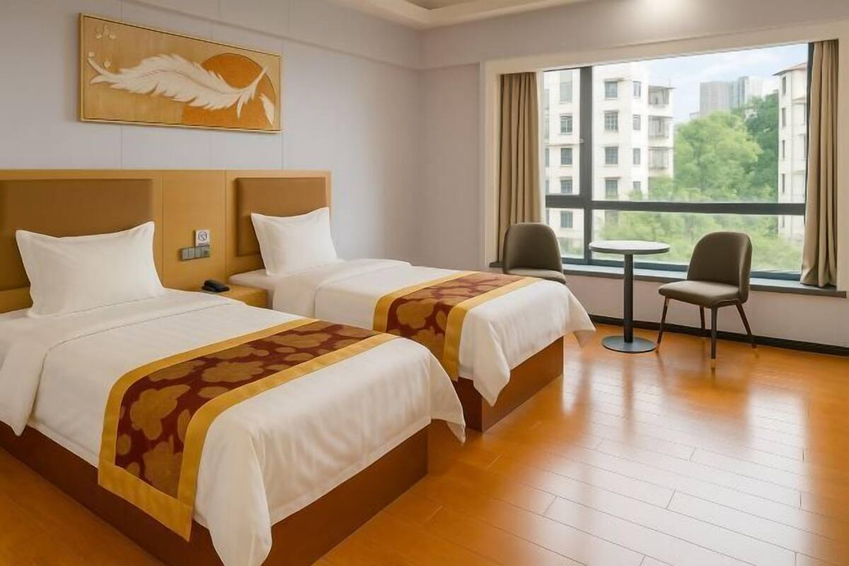 Habitación de hotel con dos camas, una mesita, una silla y ventana con vistas a edificios.
