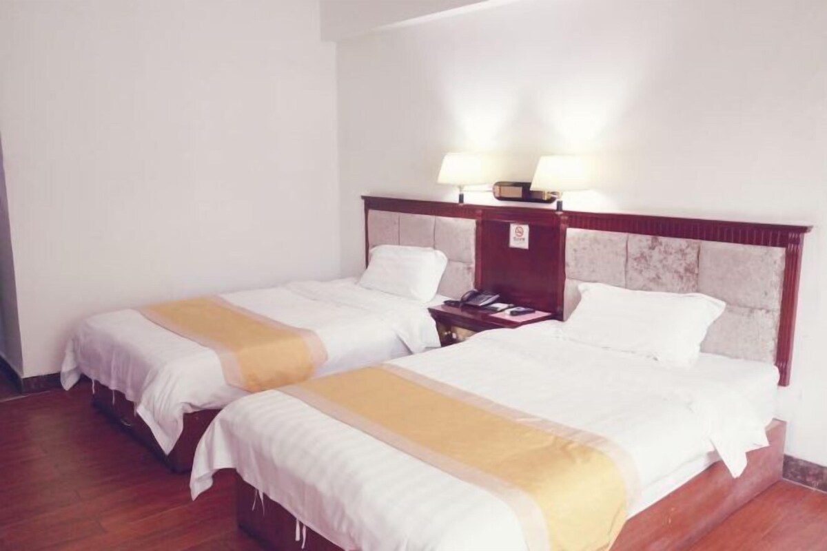 A hotel room with two single beds, wooden headboards, bedside tables with lamps, and a plain white wall.