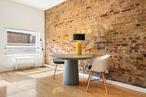 A dining area with a round table, two chairs, a yellow lamp, and a brick wall.