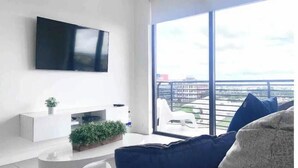 A modern living room with a flat-screen TV mounted on the wall, a white entertainment unit, and a large window offering a city view. - A705A Awesome DT Doral Apart
 (Doral)