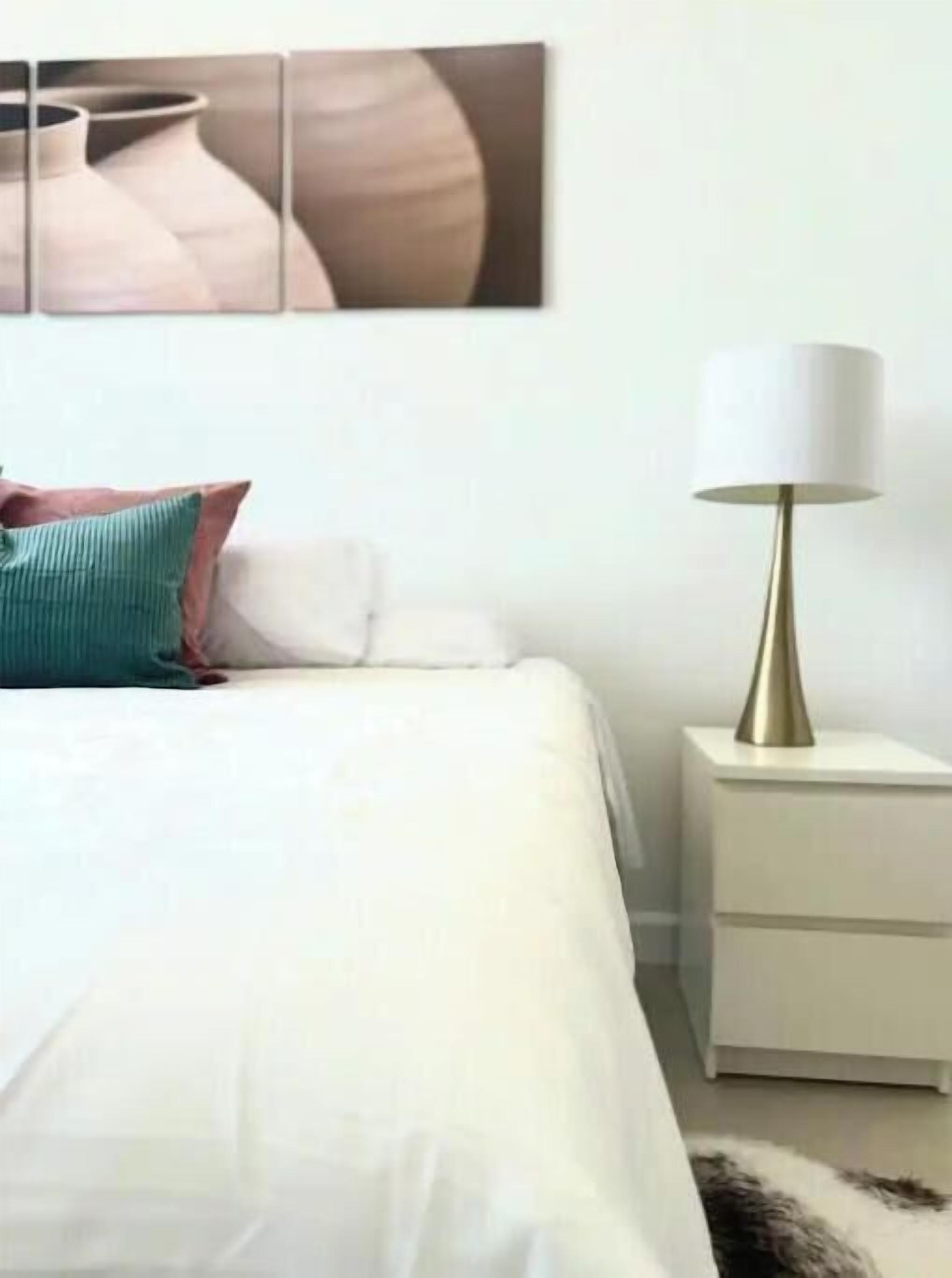 Un lit bien fait, avec une literie blanche, un oreiller aux tons turquoise et marron, et une lampe dorée posée sur une table de chevet.