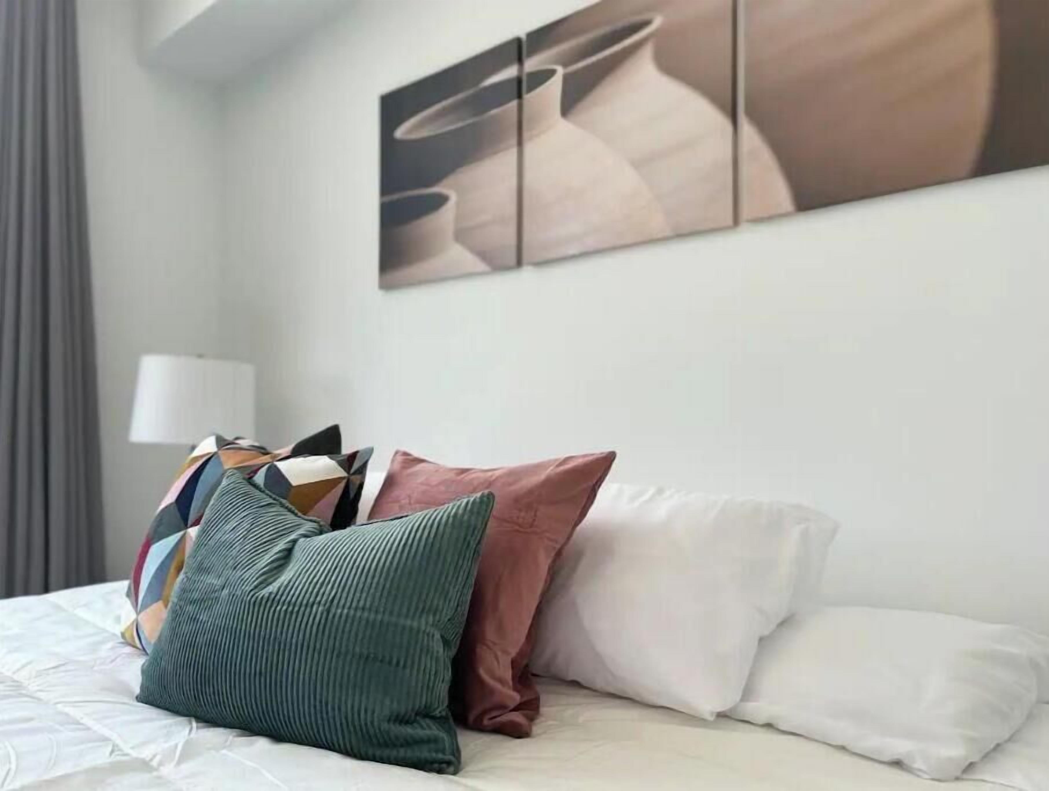 Una cama bien hecha con una colcha blanca y varias almohadas, incluyendo una verde con textura y una rosa.