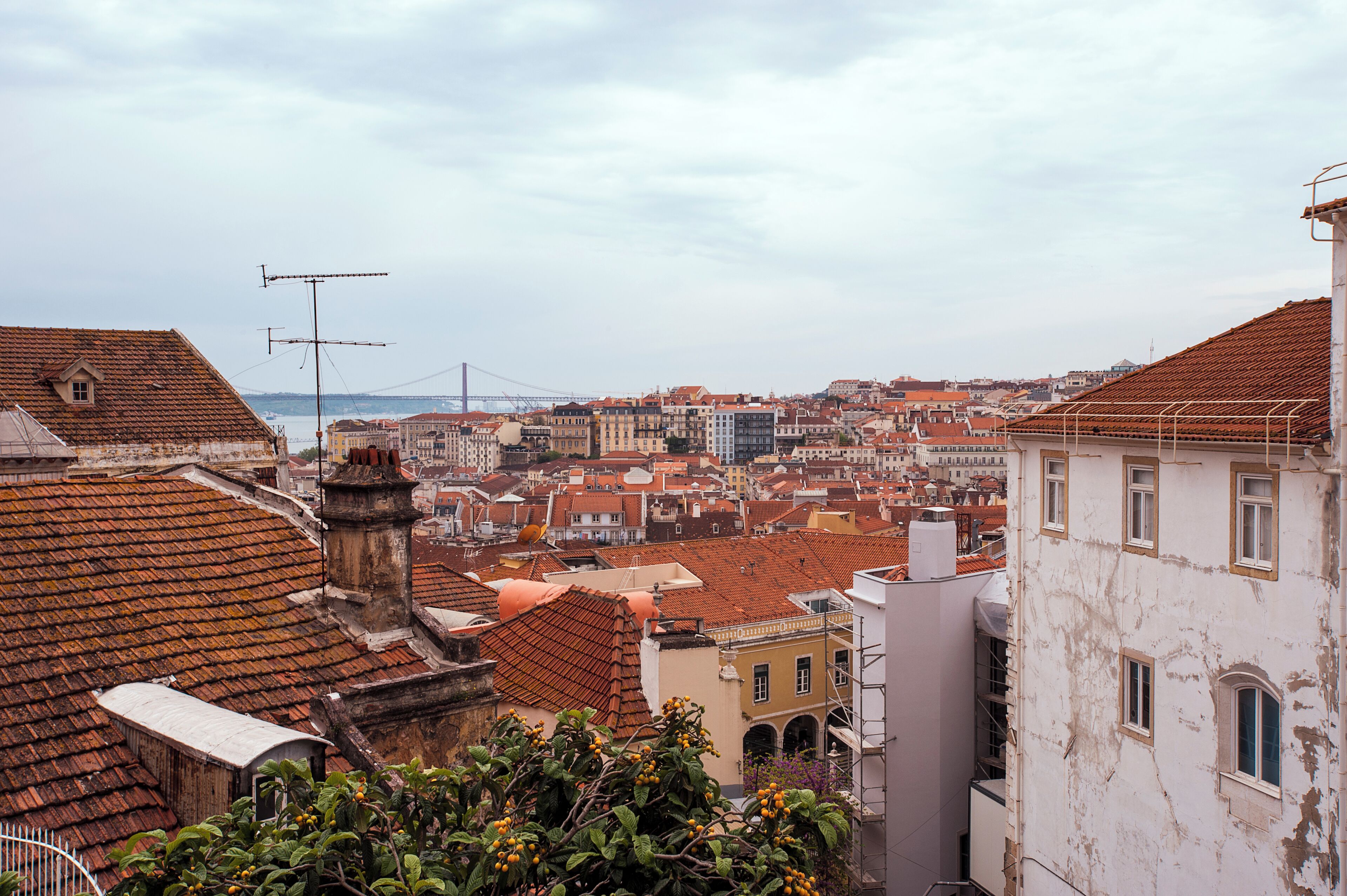 A cityscape with red-tiled rooftops, a bridge in the distance, and a mix of historic and modern buildings.