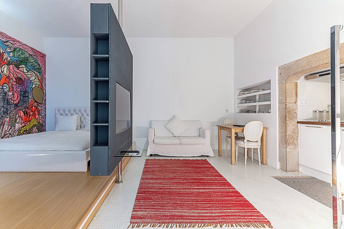 A modern living room with a bed, TV, white sofa, dining table, and kitchen area.