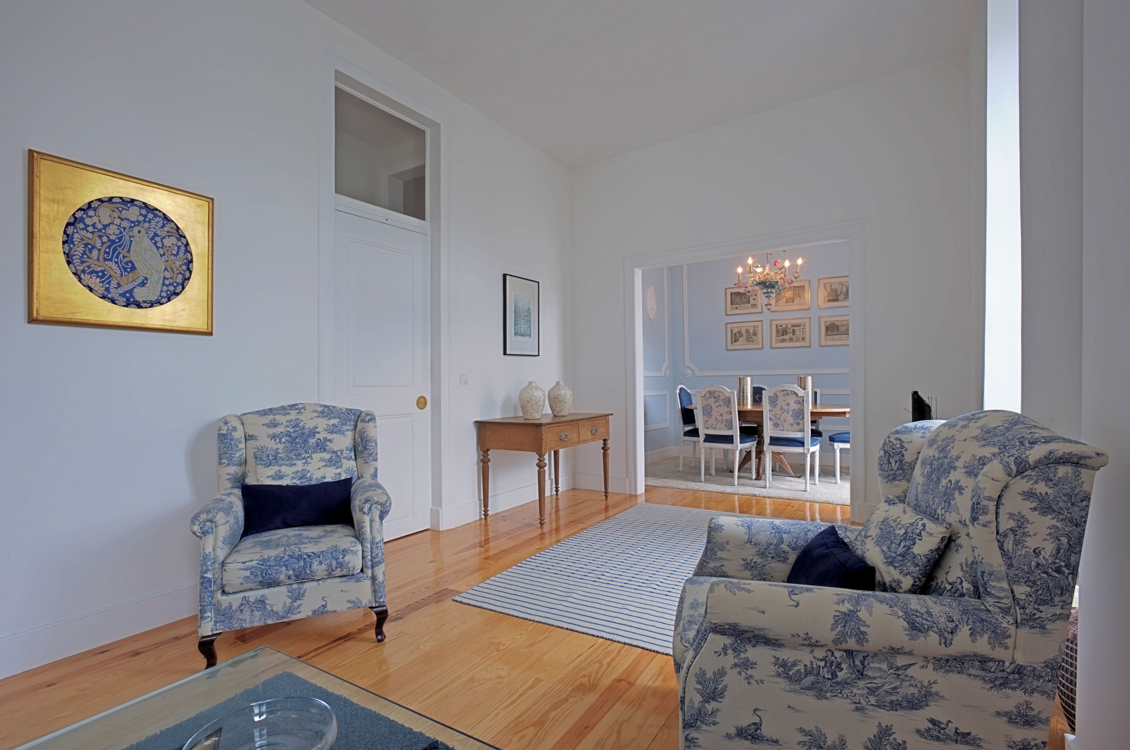 A living room with two patterned armchairs, a wooden coffee table, and a rug. There is a door leading to another room with a dining area.