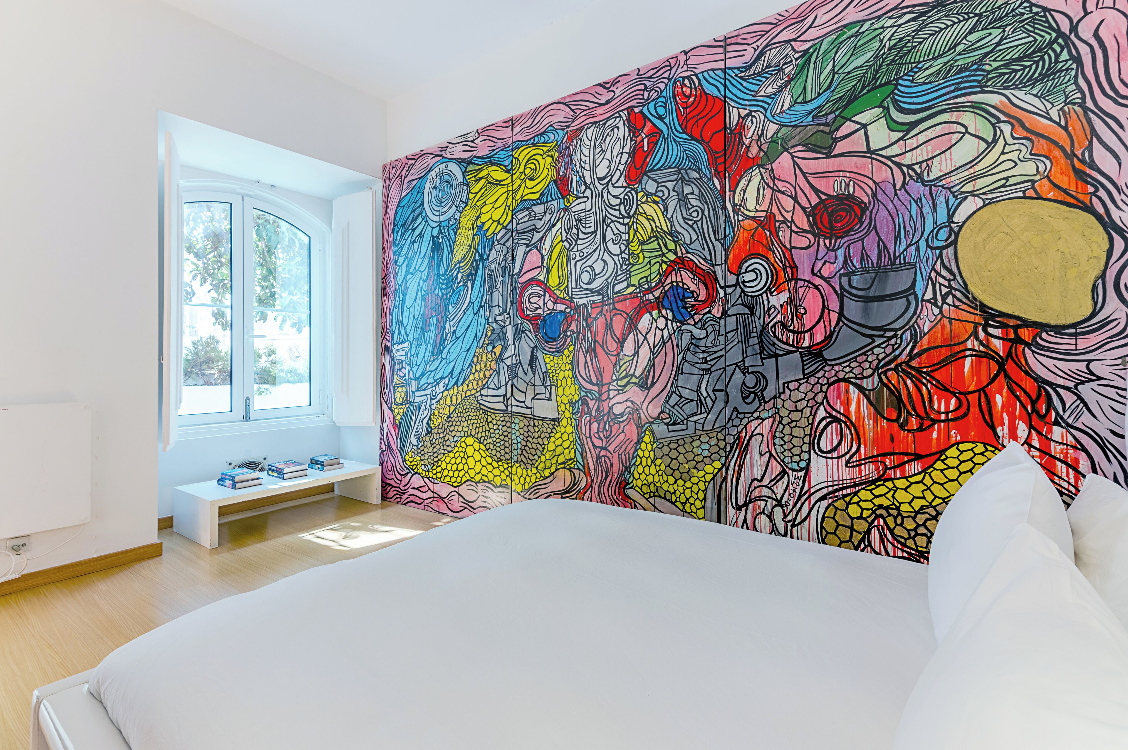 Une chambre avec une grande fresque abstraite sur le mur, une fenêtre donnant sur des arbres et un lit blanc.