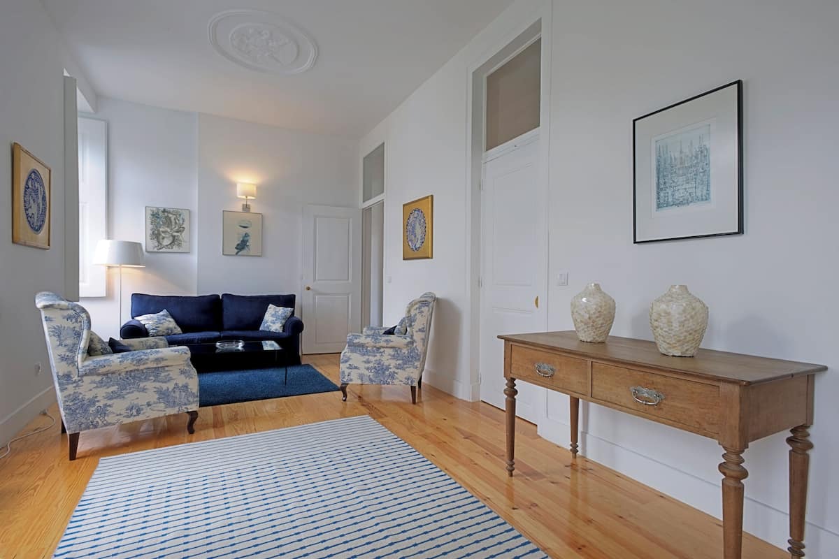 A living room with a blue sofa, two patterned armchairs, a wooden coffee table, and a blue and white striped rug.