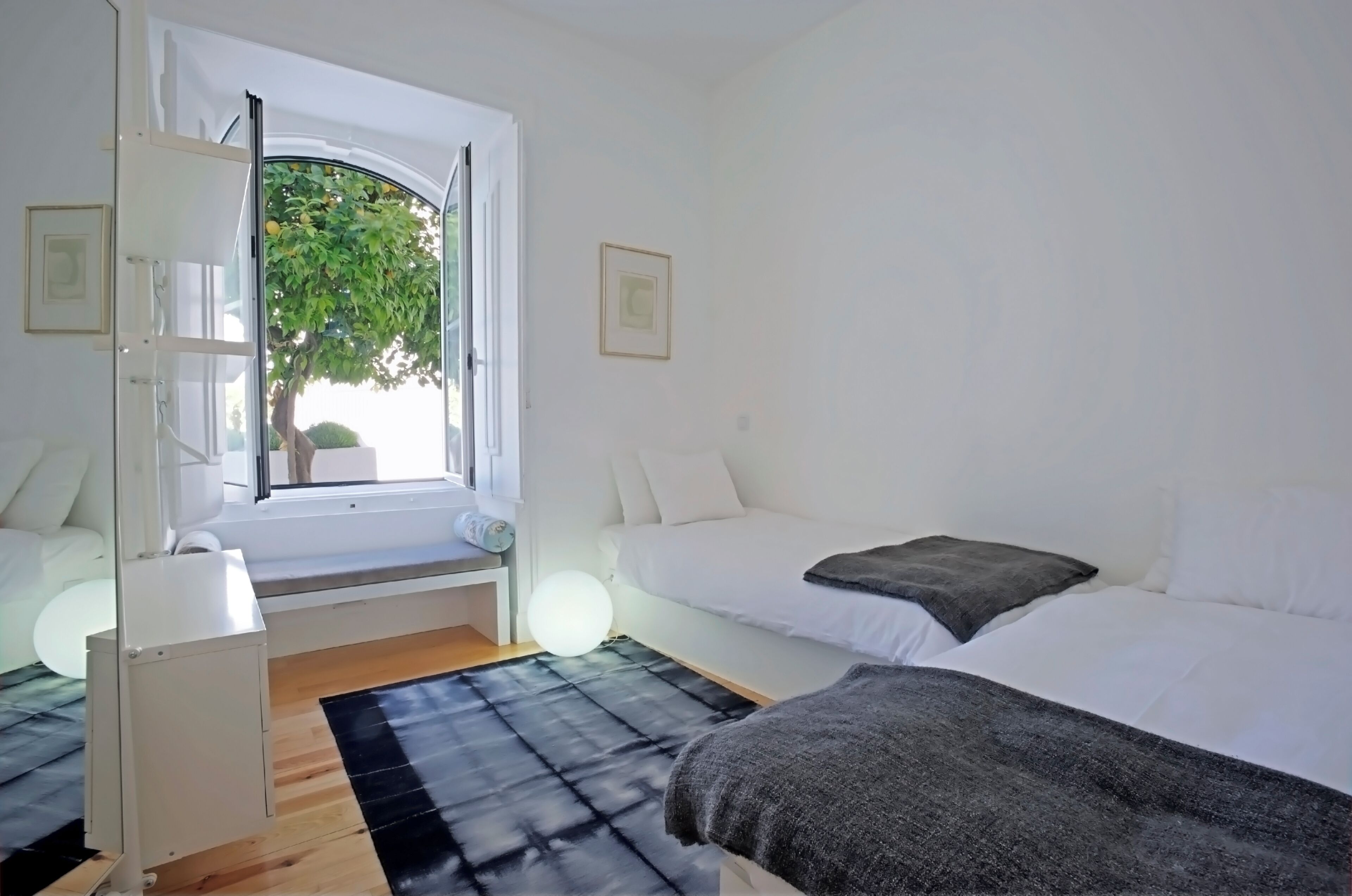 A modern bedroom with a large window, a bed, a bedside table, and a small cabinet.