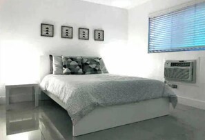 A modern bedroom with a large bed, a bedside table, and a wall-mounted air conditioner. - Cozy 1/1 Apartment in the heart of Coconut Grove (Miami)