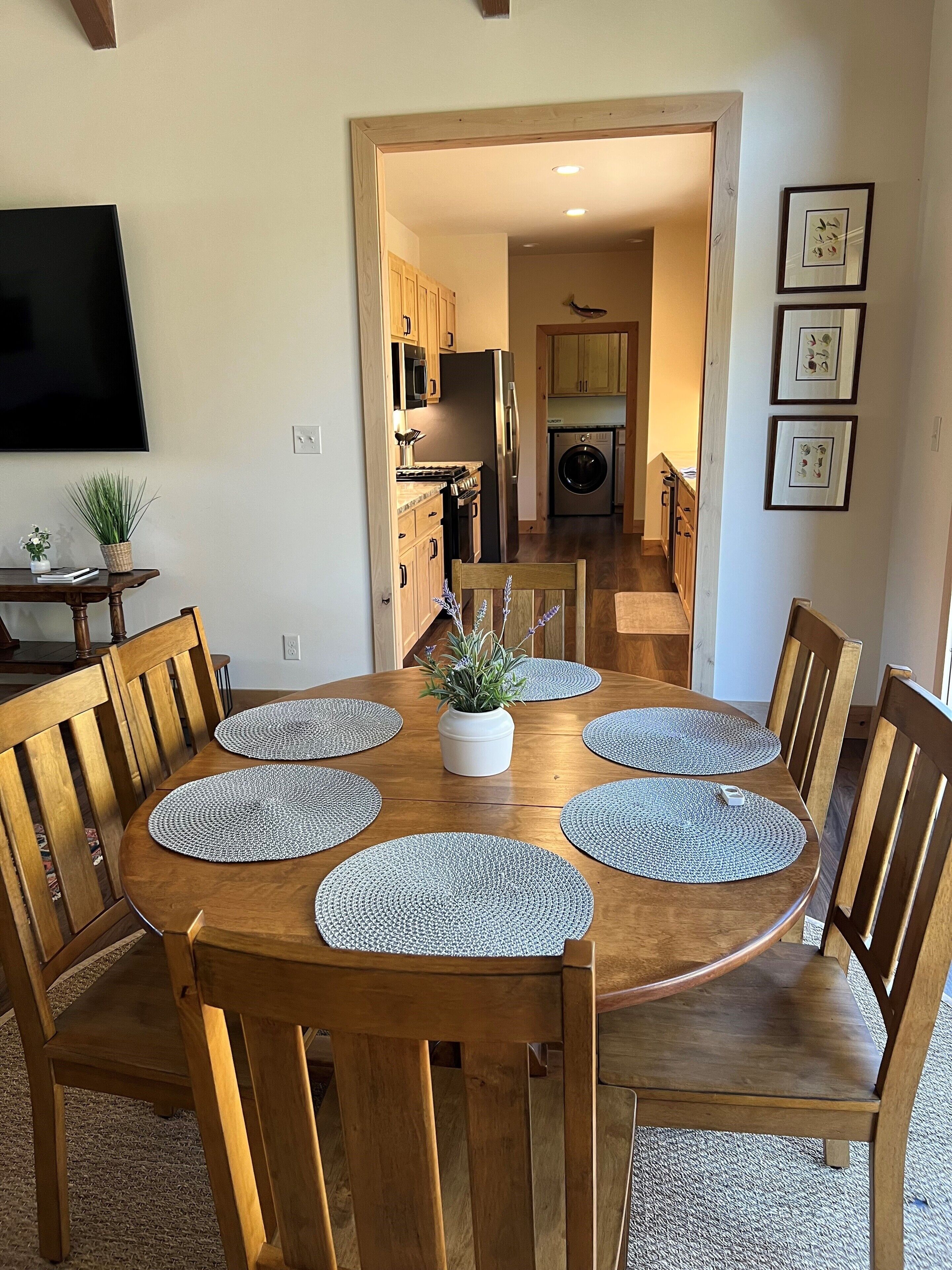 Un coin repas avec une table ronde en bois, six chaises et des sets de table. Une plante en pot est posée sur la table.