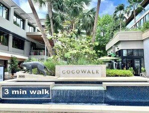 Señal de 'Cocowalk' con el rótulo '3 minutos a pie', rodeada por una fuente y palmeras.