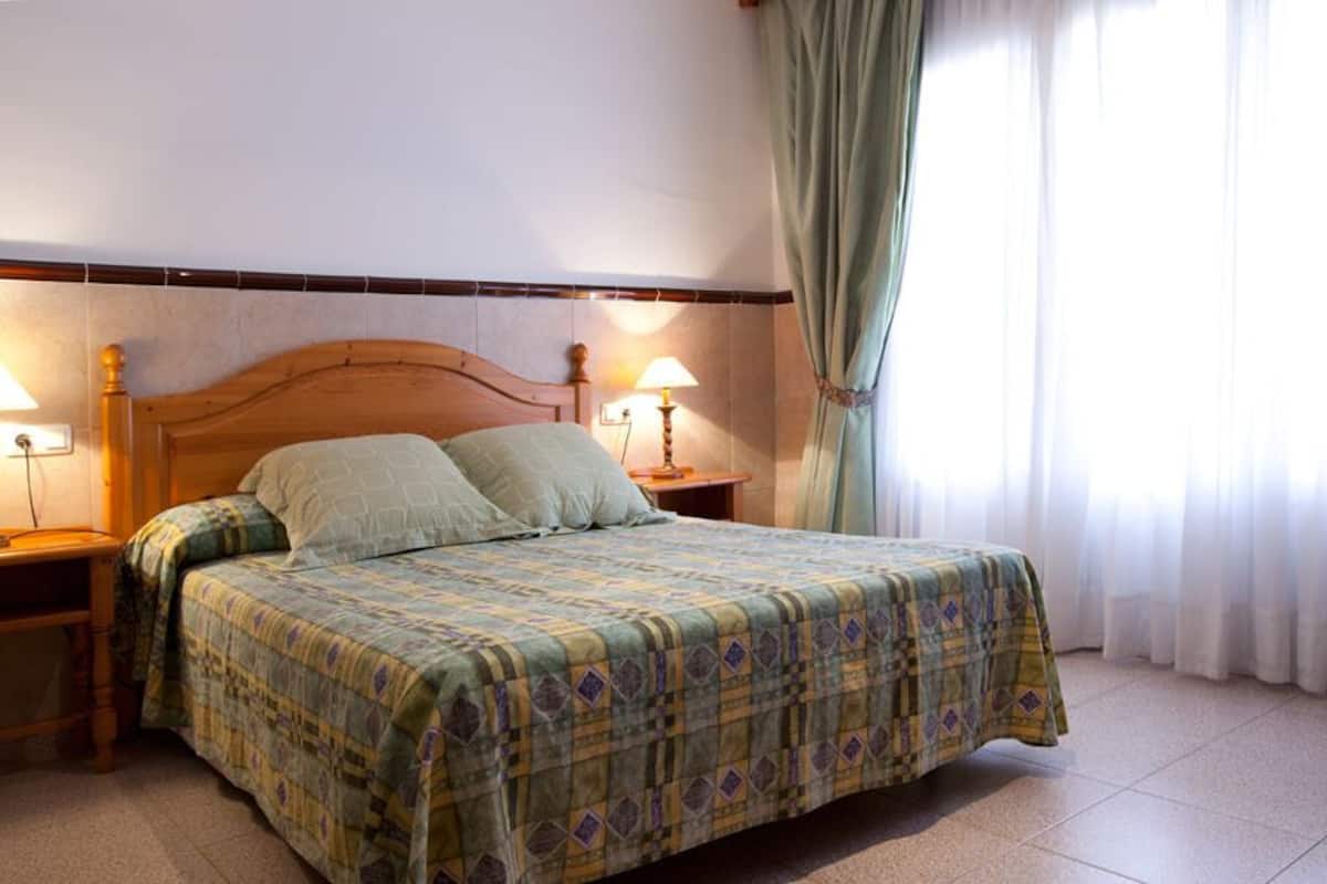 A bedroom with a wooden headboard, a bed with a patterned bedspread, two bedside lamps, and a window with sheer curtains.