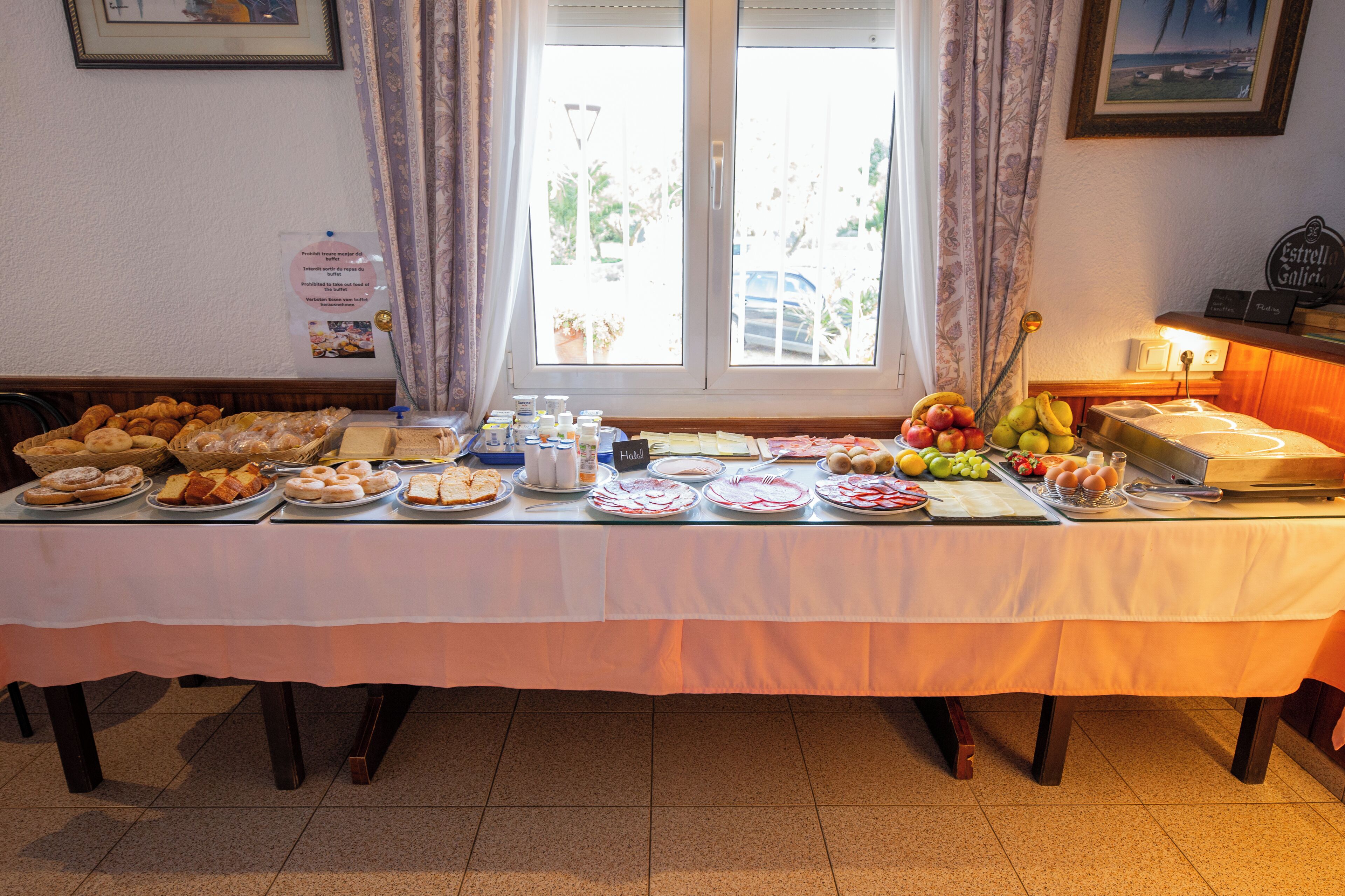 A breakfast buffet table with various food items and a window with curtains.
