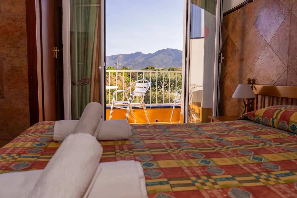 A hotel room with a bed, a balcony with chairs, and a mountain view.