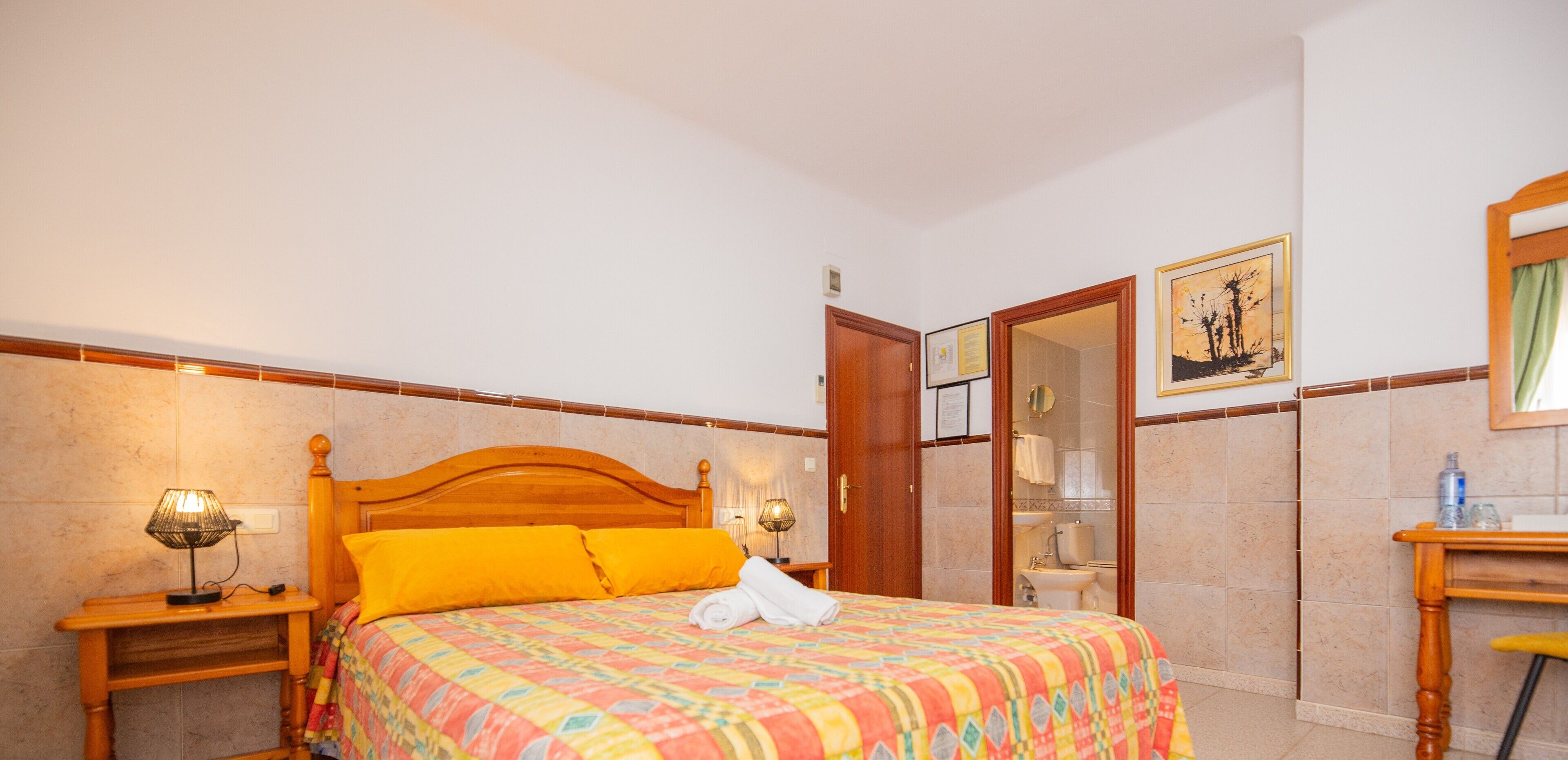 A hotel room with a wooden bed, a nightstand with a lamp, a bathroom visible through an open door, and a framed picture on the wall.
