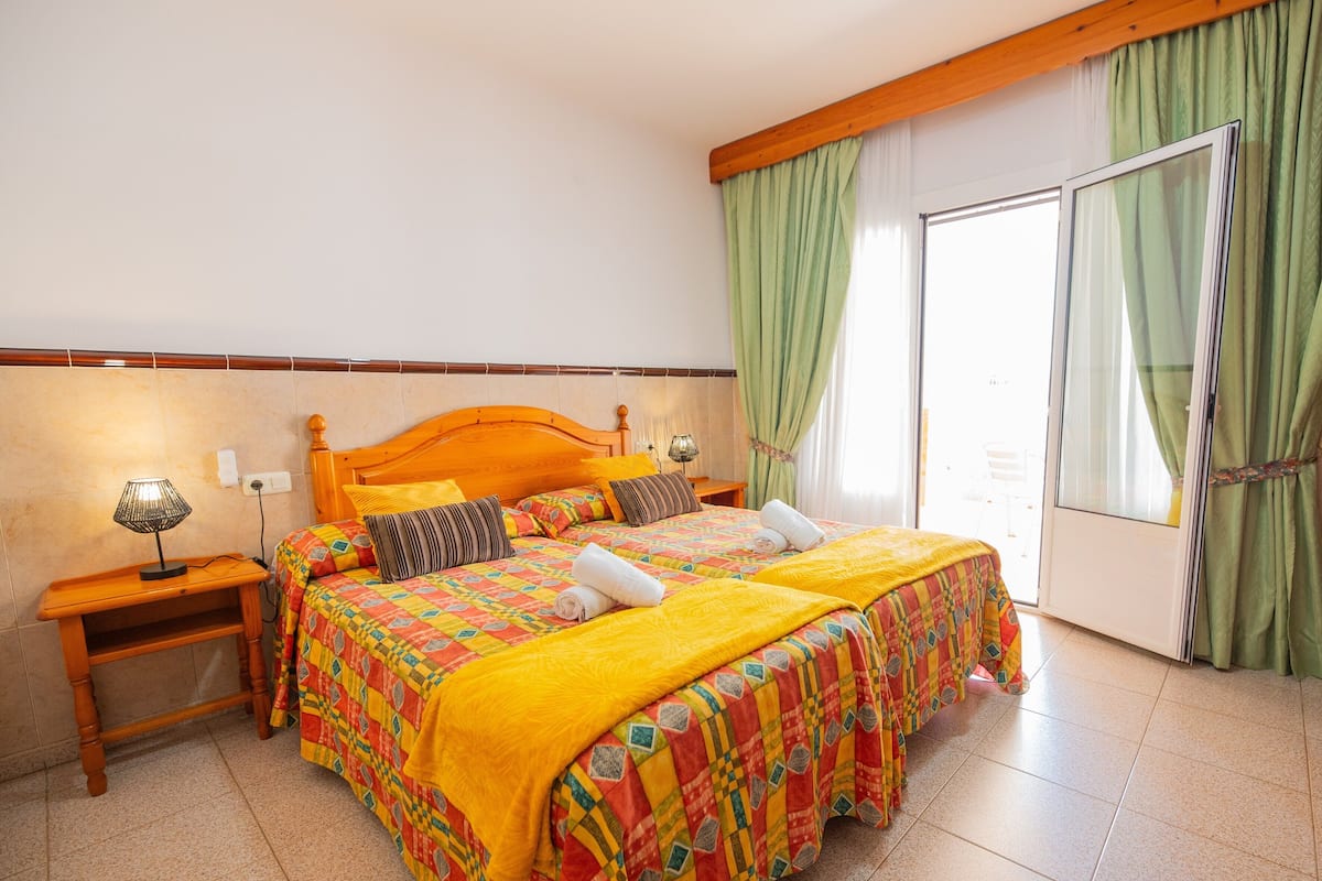A double bed with a colorful quilt, a nightstand with a lamp, and a view of a green outdoor area through a sliding door.