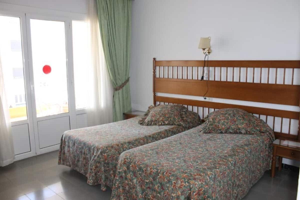 A room with two single beds, a wooden headboard, a nightstand, a lamp, and a window with curtains.