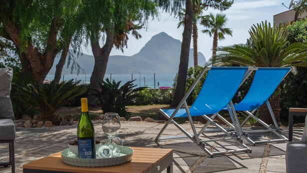 A patio with two blue lounge chairs, a table with a bottle and glass, and a view of the sea and mountains. - Villa Makar Sea View (San Vito Lo Capo)
