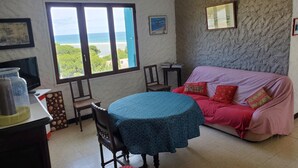 Un salon chaleureux avec une grande fenêtre donnant sur la mer, une table ronde entourée de chaises, un canapé agrémenté de coussins à motifs et un lustre.