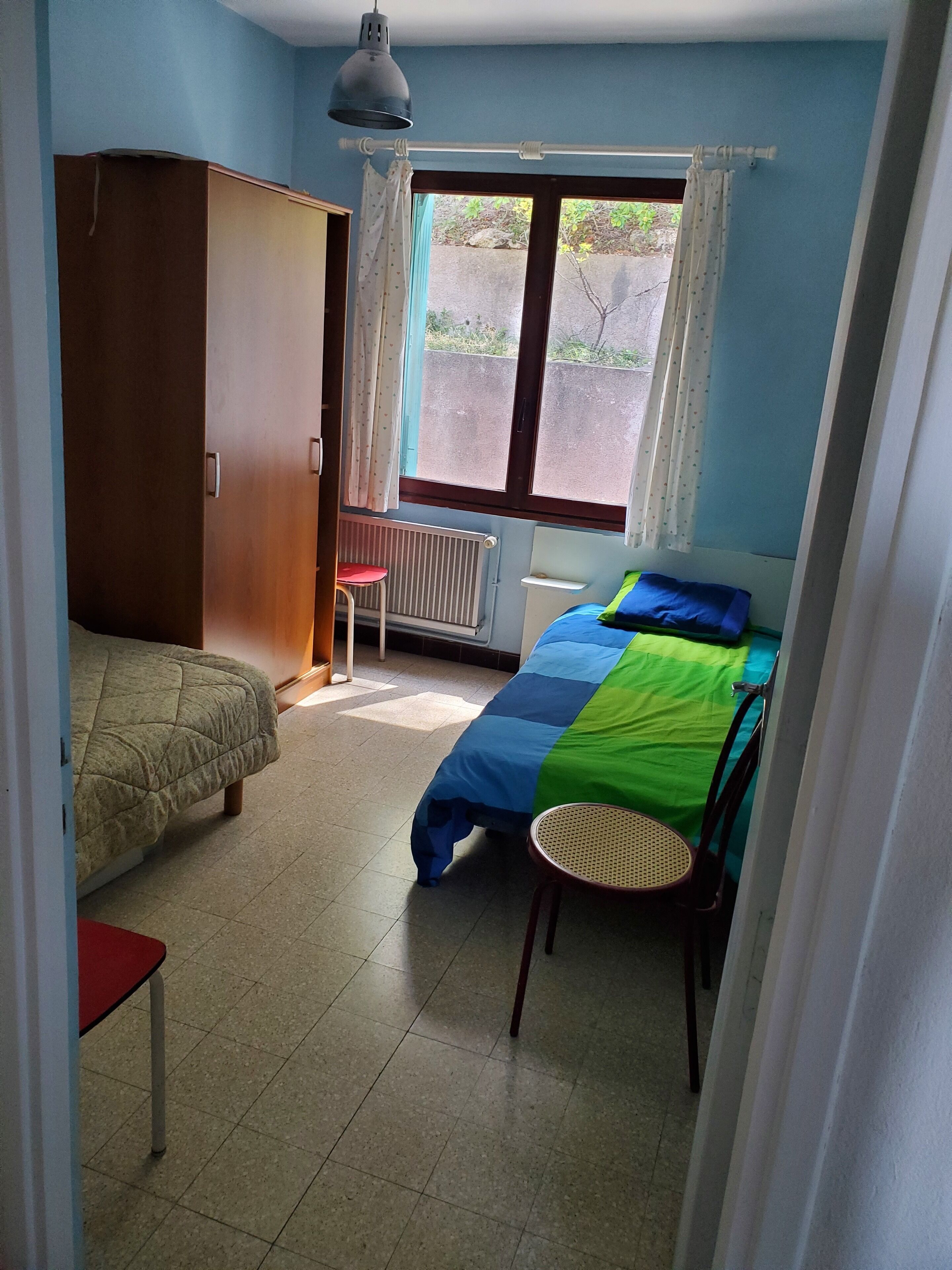 Une chambre à coucher avec un lit, une armoire, une chaise, une table et une fenêtre avec des rideaux.