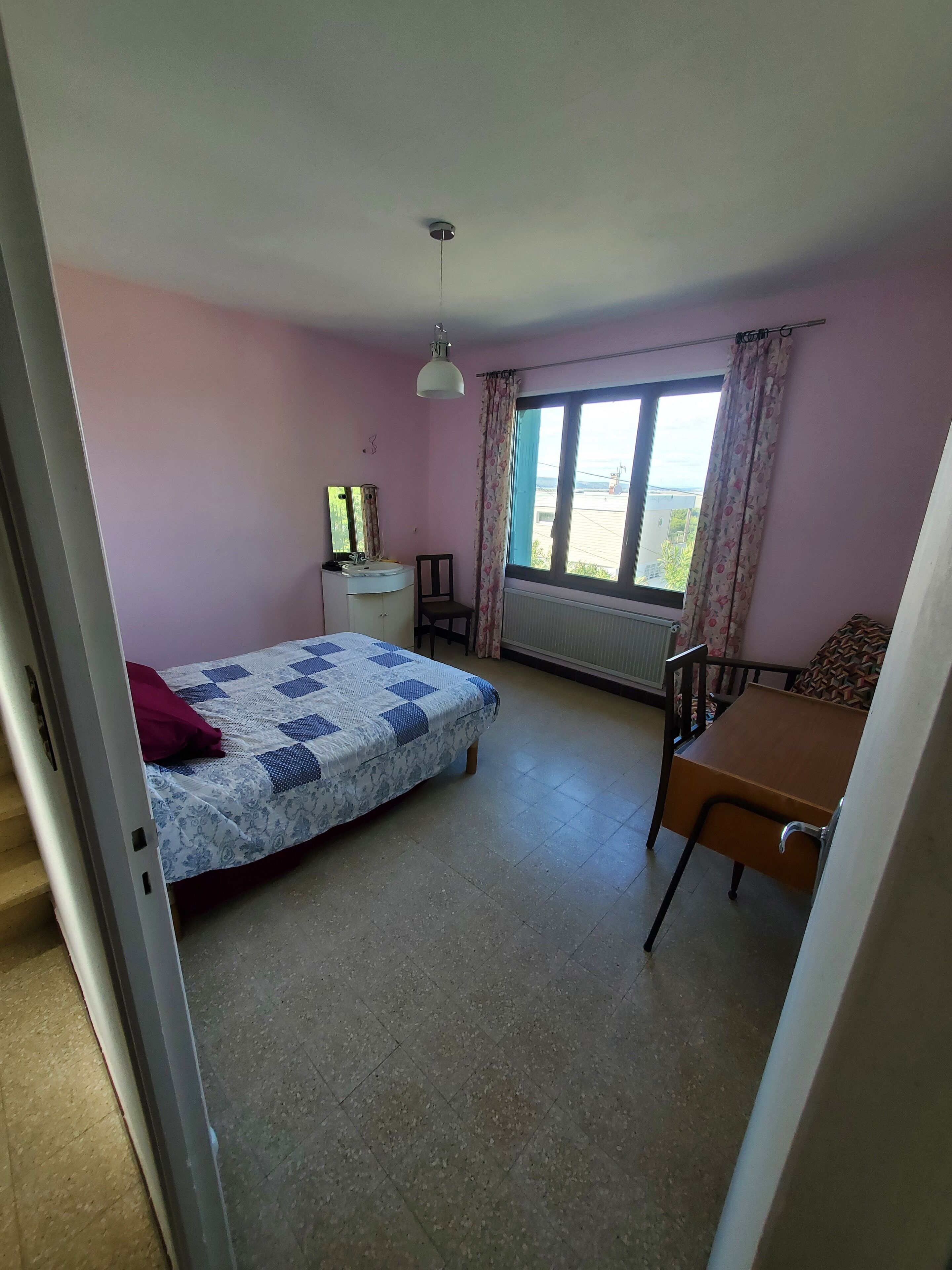 A bedroom with a bed, a desk, a chair, a window with curtains, a radiator, and a ceiling light.