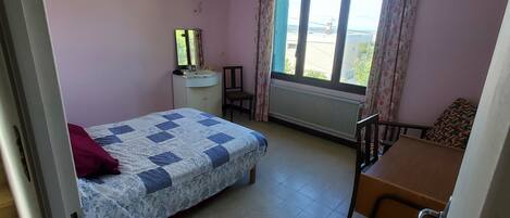 A bedroom with a bed, a desk, a chair, a window with curtains, a radiator, and a ceiling light.