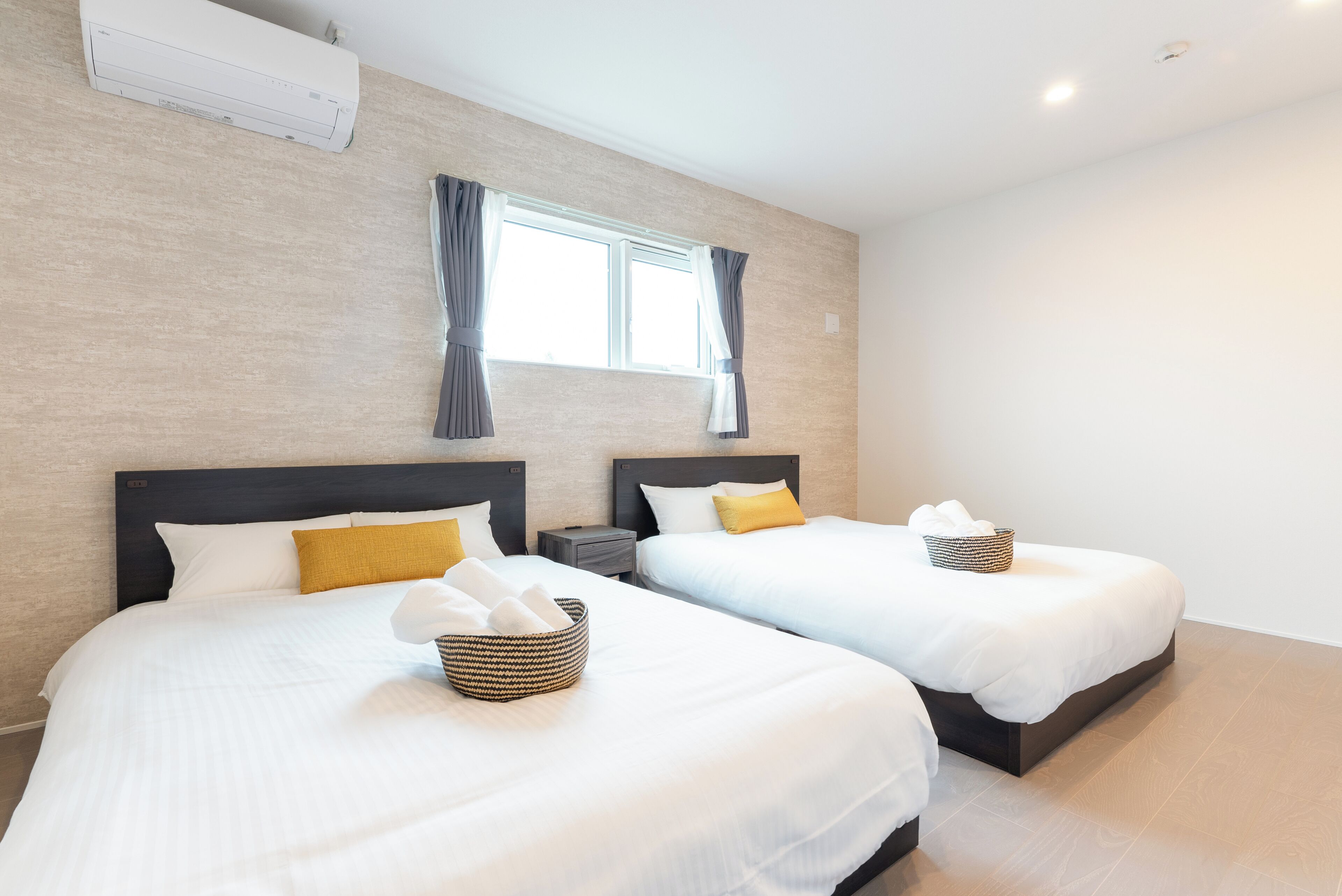 A hotel room with two beds, each with white bedding and a yellow pillow. There is a window with curtains, a bedside table, and a wall-mounted air conditioner.