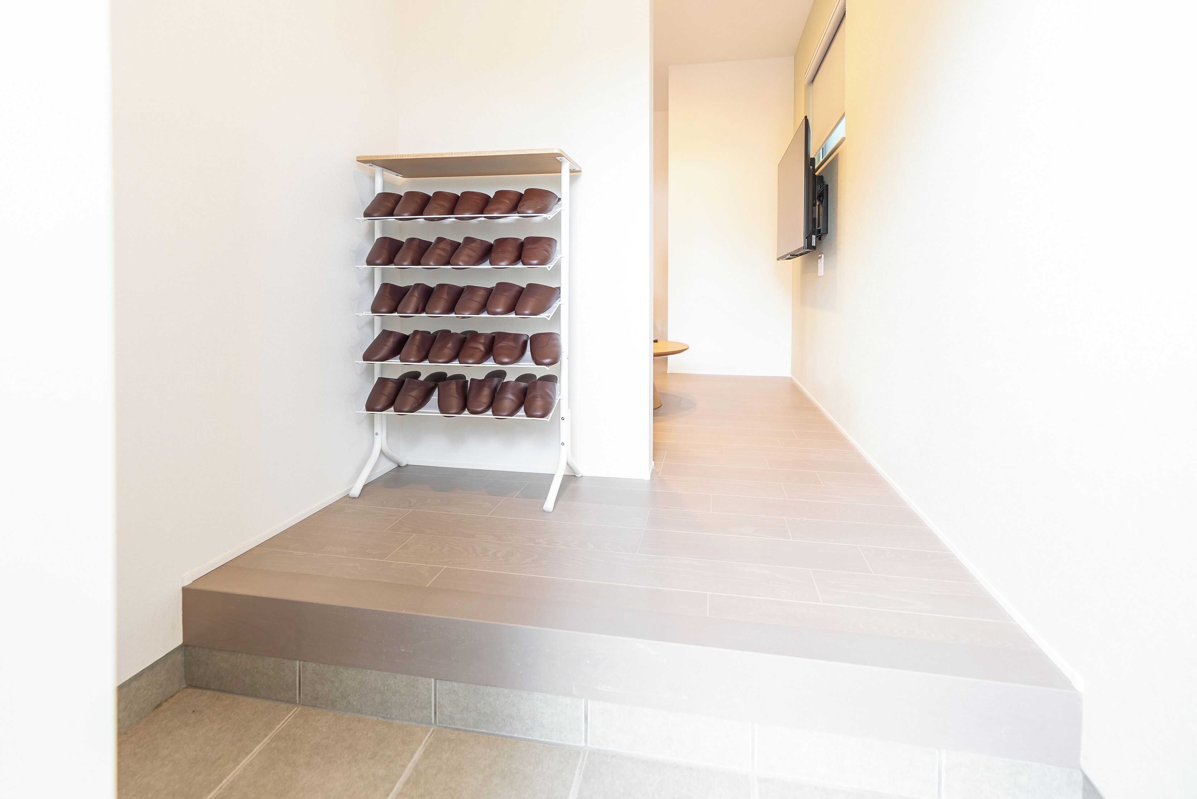 A shoe rack with multiple pairs of shoes against a wall in a hallway with a tiled floor.